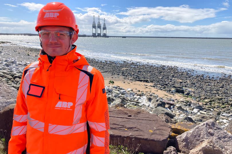 A man in protective clothing stands near the coast