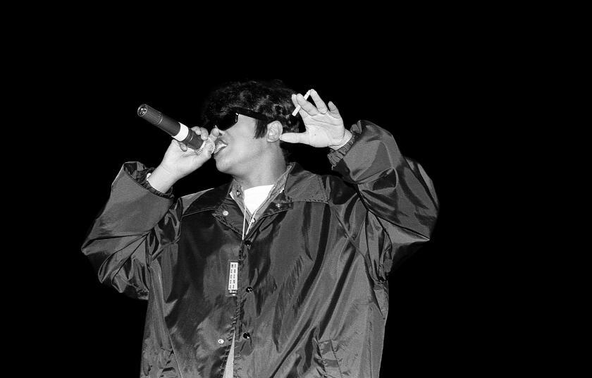 A blanck and white photo of a woman in a large black jacket standing on a stage and speaking into a microphone