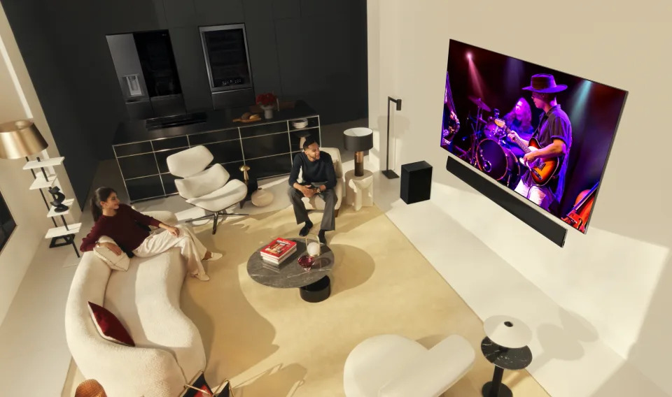 Some people in a nice white living room watching live music on TV.