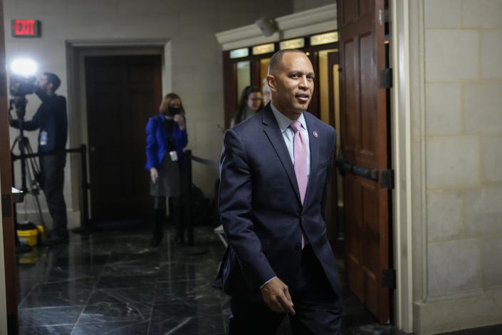 Rep. Hakeem Jeffries passes from a corridor in the Capitol through a wooden door, as members of the media gather behind him, one adjusting a portable spotlight. 