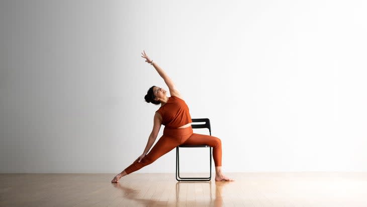 A woman with dark hair practices Reverse Warrior on a chair. She is wearing copper colored tights and top .