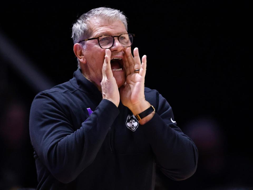 geno auriemma man wearing glasses navy pullover holds hands up to mouth to yell against dark background