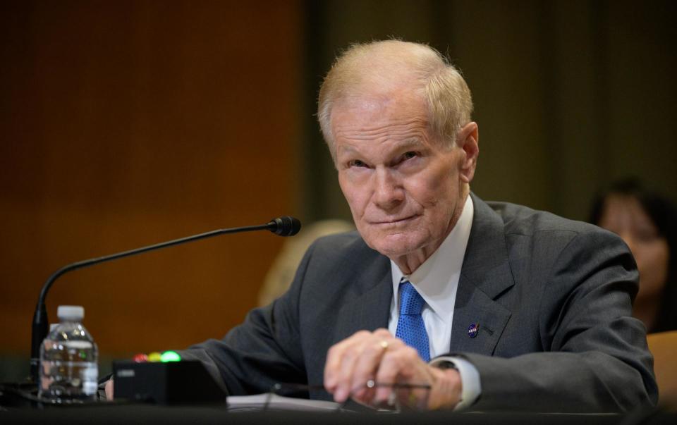 a man in a suit sits at a table covered in microphones in a large wood-paneled room