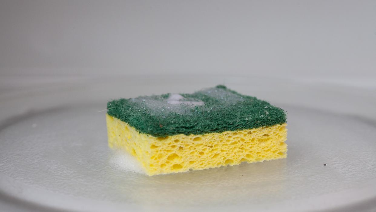 A green and yellow sponge covered in soap suds sits inside of a microwave.