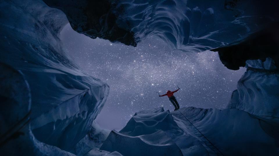 From the interior of a glacier, a person standing on the edge and the night sky are visible.