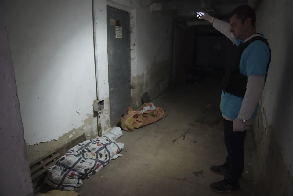 A police officer shines a light at what appear to be two lifeless bodies beneath blankets.