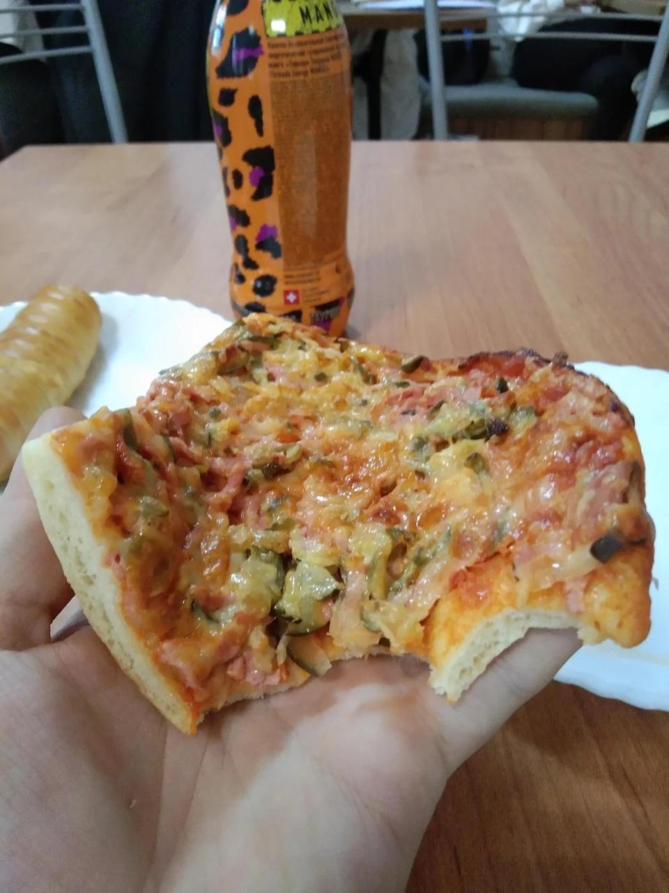 A hand holding a slice of pizza with visible toppings, including cheese, vegetables, and possibly meat, with a bottle of beverage and a loaf of bread in the background