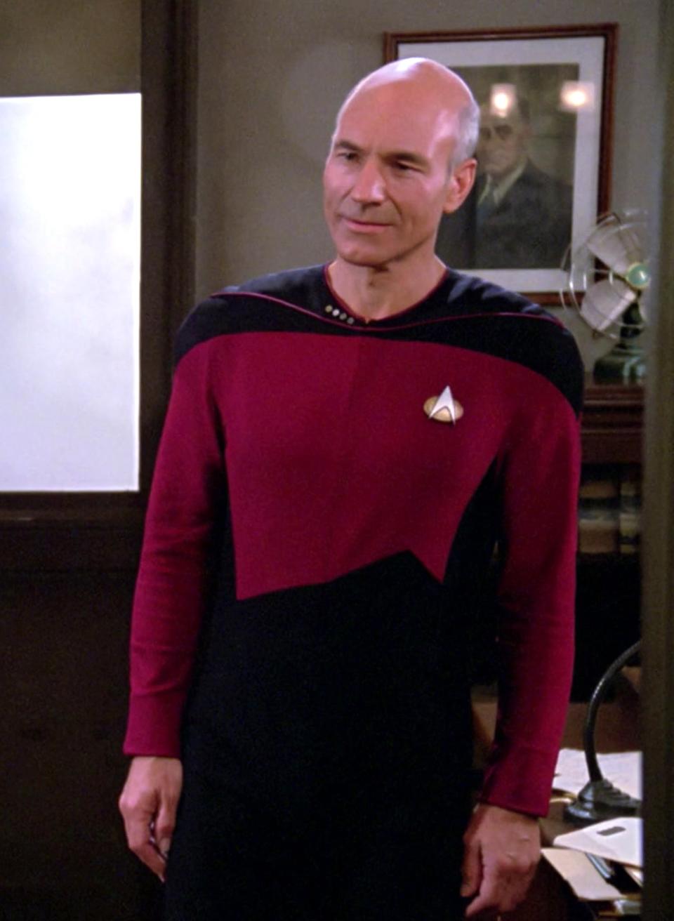 Patrick Stewart dressed as Captain Jean-Luc Picard in a dark uniform stands next to a door