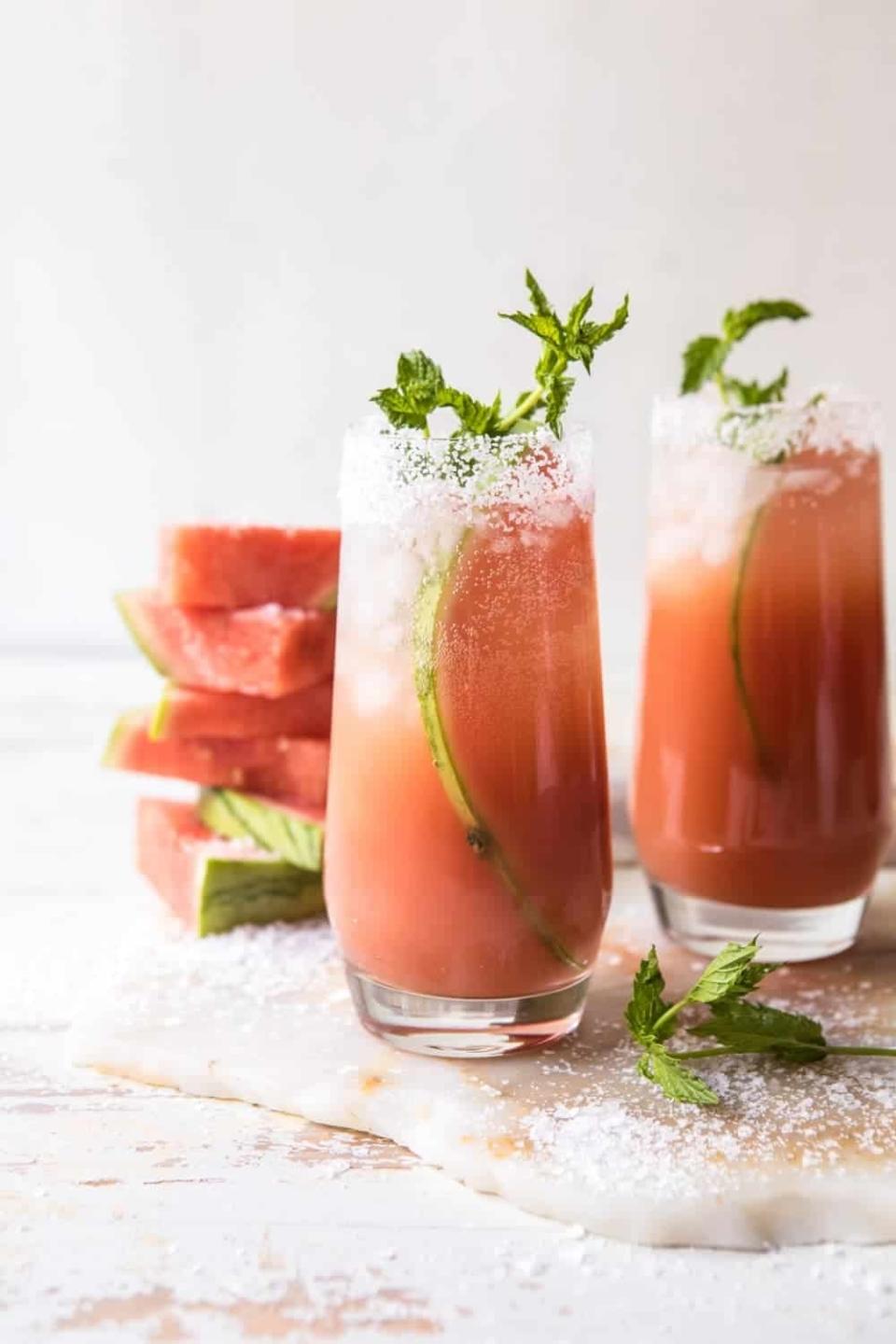 Two glasses of a watermelon paloma garnished with mint and a salted rim, accompanied by watermelon slices in the background