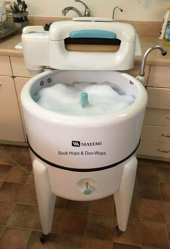 A retro-styled Maytag washing machine labeled "Sock Hops & Doo-Wops" with foamy water inside, situated in a kitchen environment