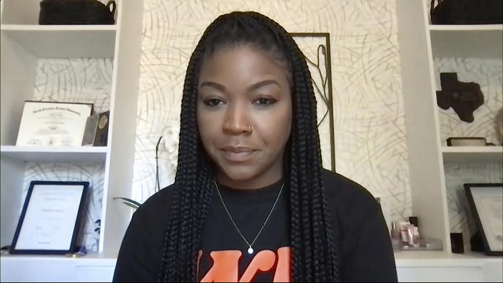 Cherelle, talking in front of a bookcase that contains framed certificates and an outline of Texas, pauses, with a look of pursed frustration expression on her face.