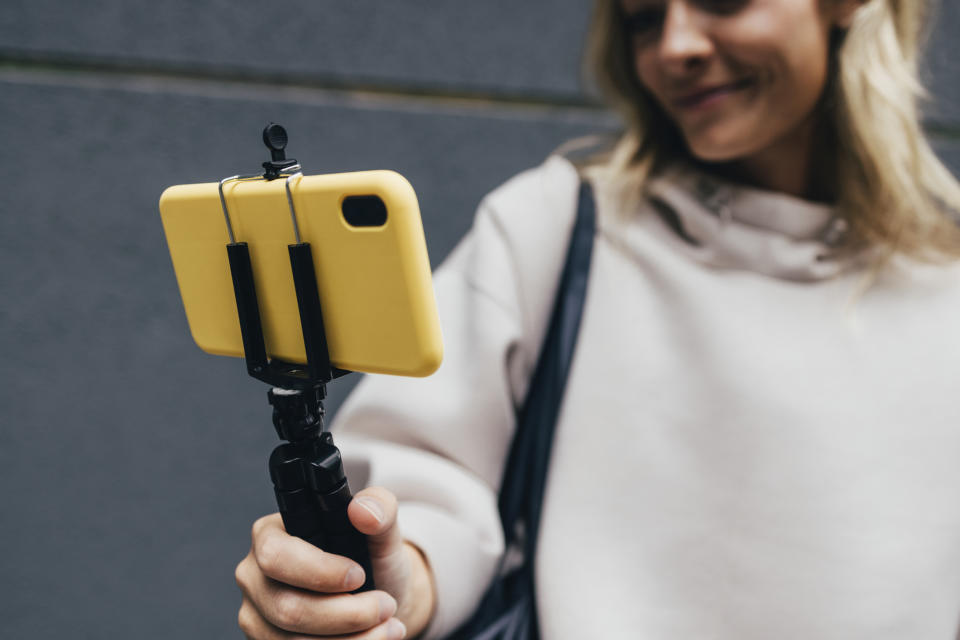 A person smiles as they hold a smartphone mounted on a tripod selfie stick, suggesting they are taking a photo or recording a video
