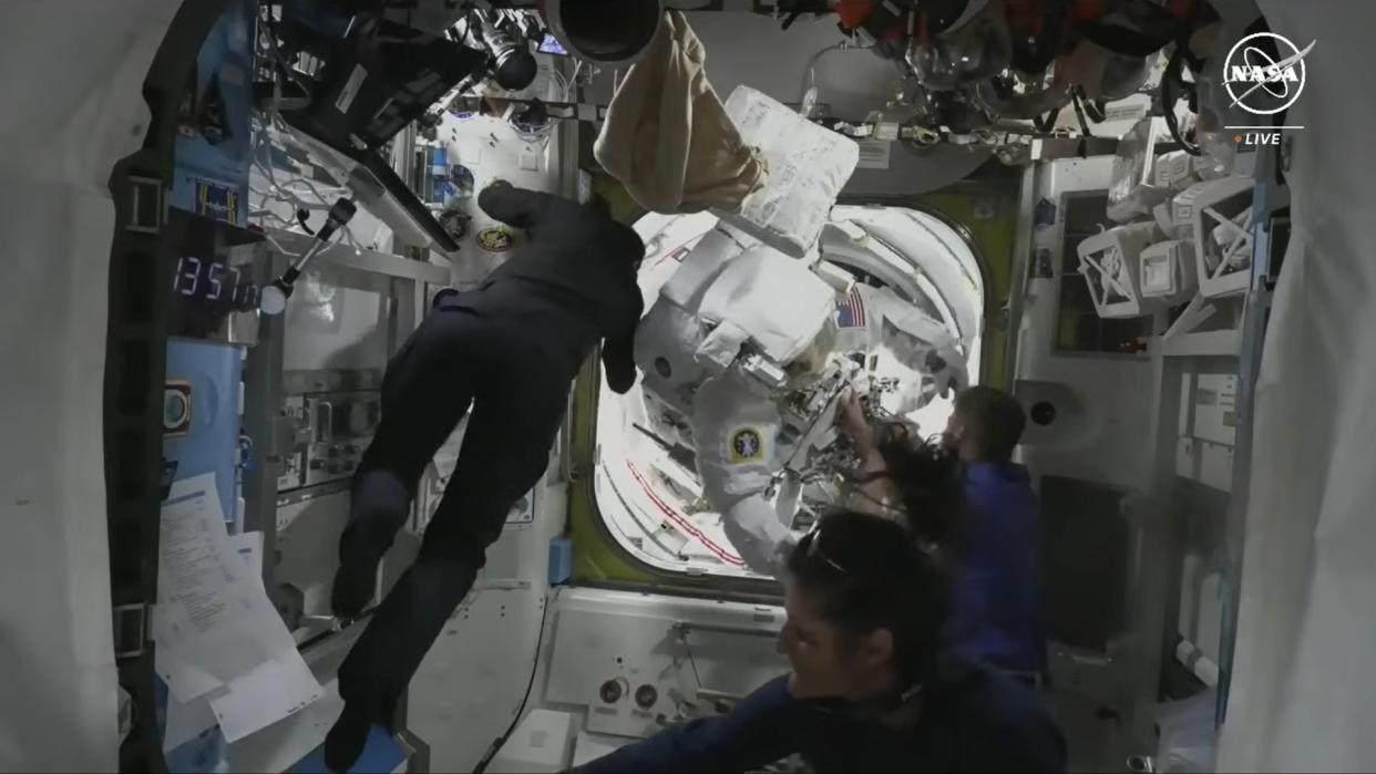  An astronaut in a bulky white spacesuit is brought into a cramped laboratory from an airlock. 