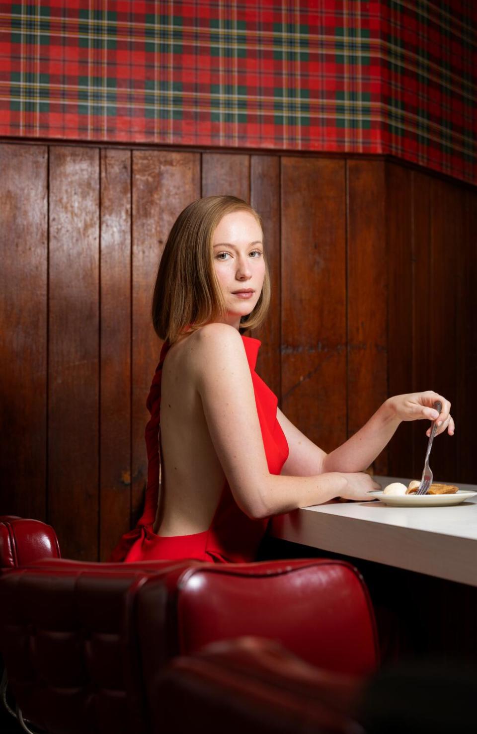 Hannah Einbinder in a backless red dress sits at a diner counter sticking a fork into something on her plate