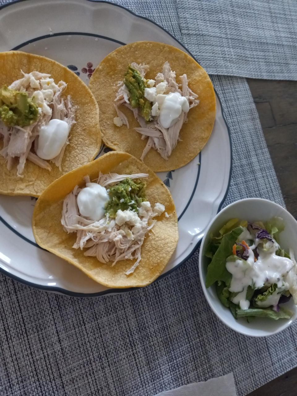 Three chicken tacos with toppings on a plate next to a small salad bowl