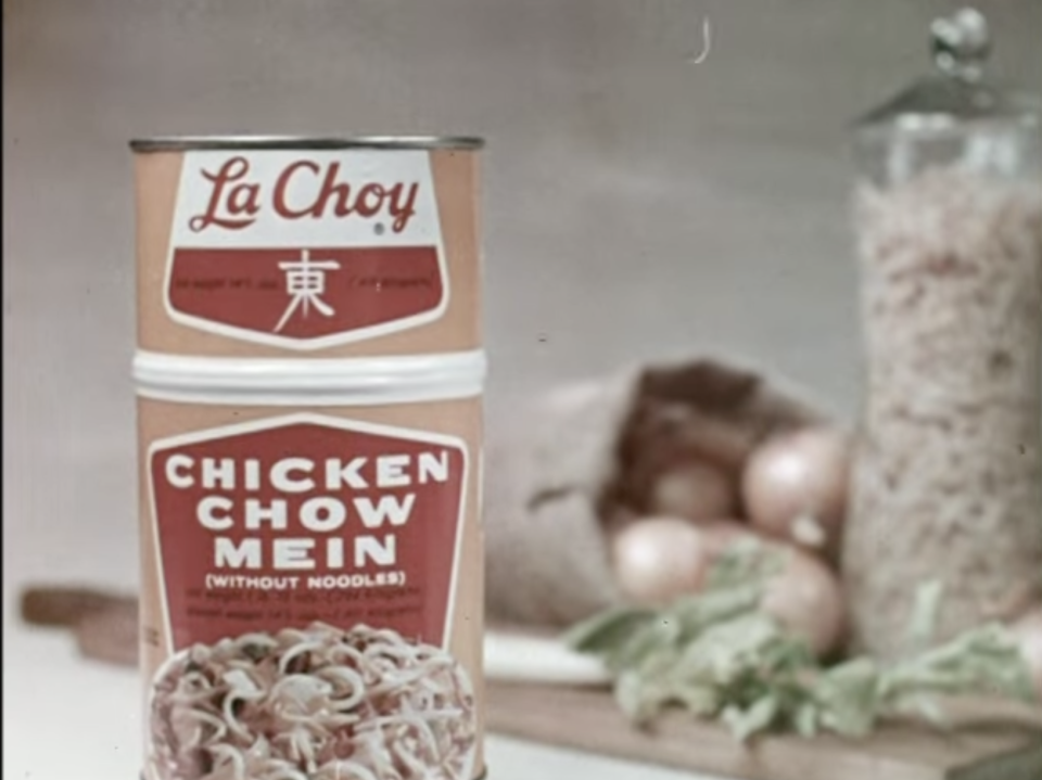 La Choy Chicken Chow Mein tin can with noodles depicted on its label. Background includes onions, spilled rice, and greens on a cutting board