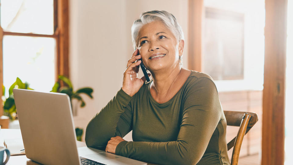 Shot of a mature woman talking on a cellphone while working on a laptop at home.