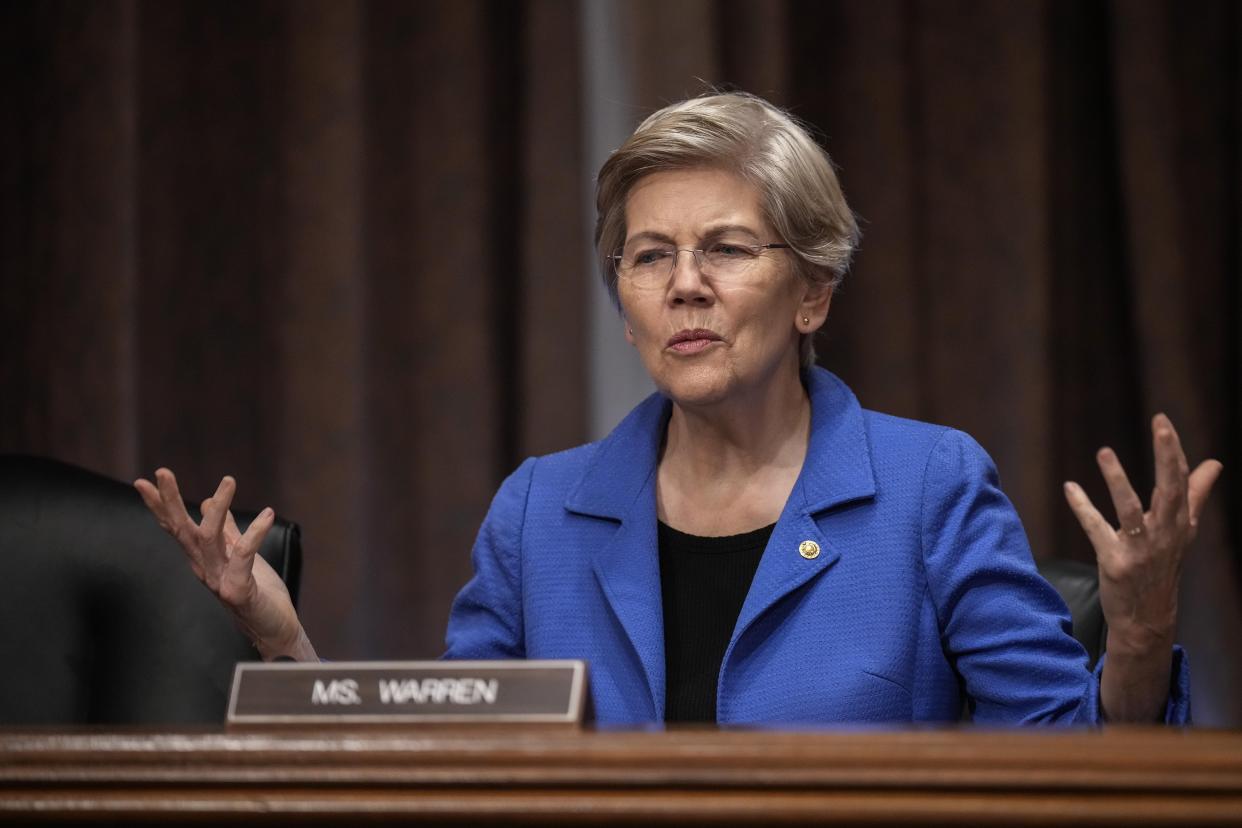 Sen. Elizabeth Warren (D-MA) in a blue jacket sitting at her seat in the U.S. Senate, holding her hands up.