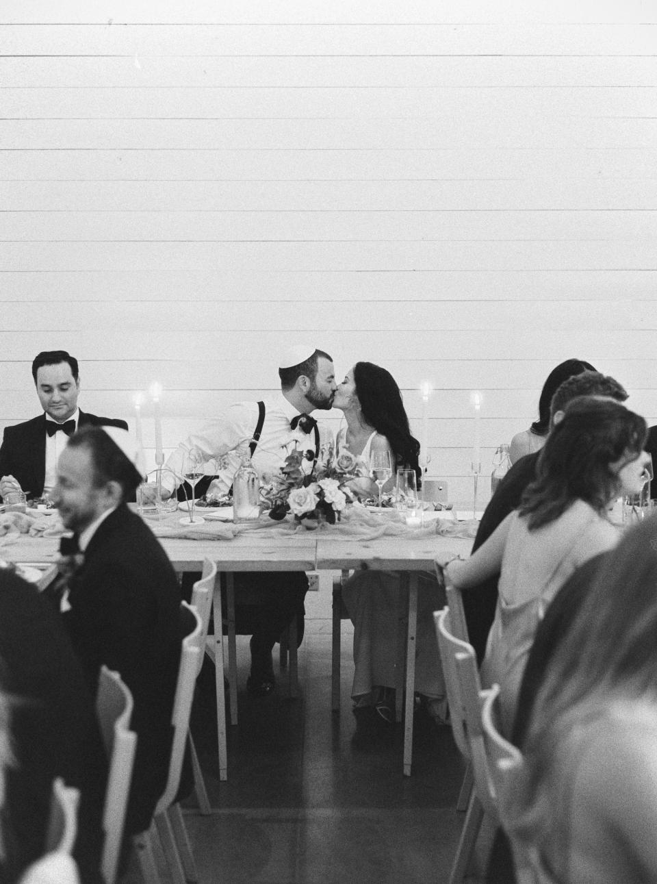 A bride and groom kiss as their wedding guests sit around them in a black and white photo.