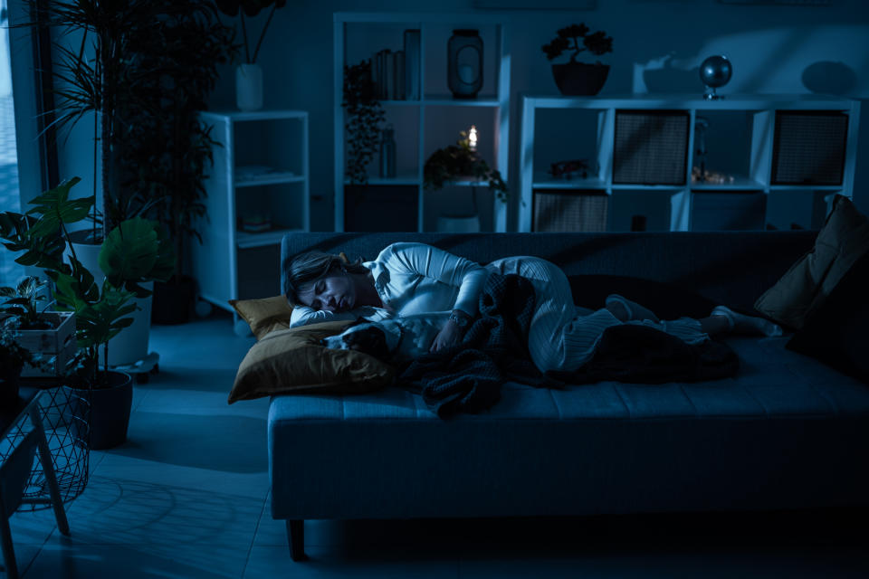 Person sleeping on a couch in a dimly lit room filled with plants and shelves, covered by a blanket