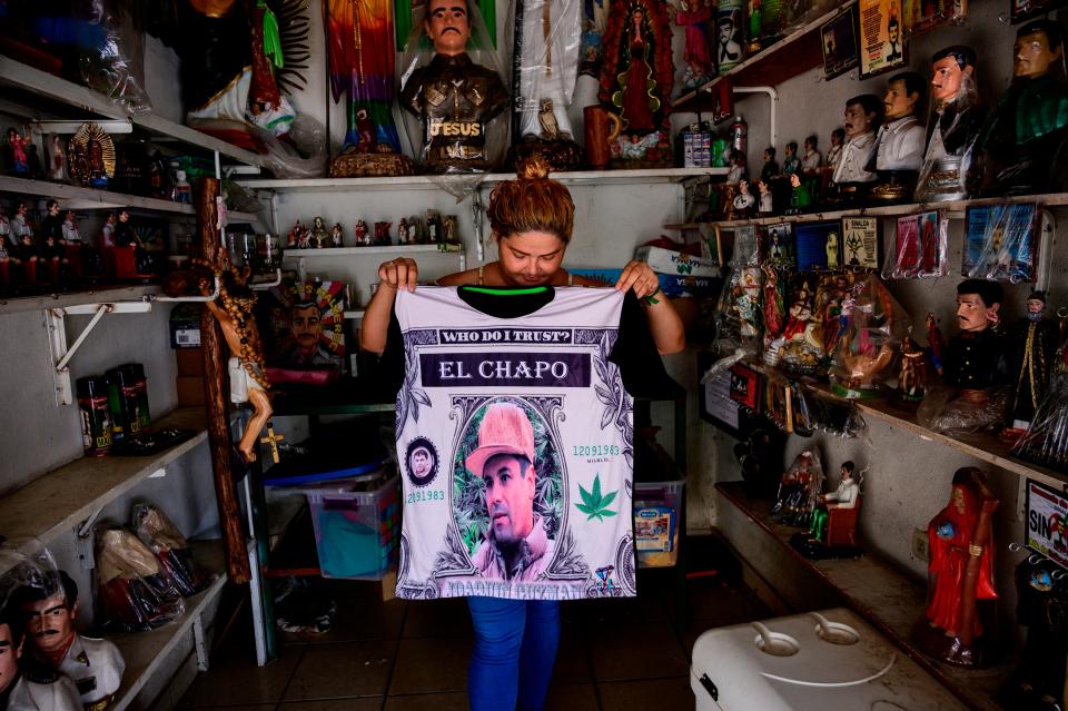 A woman in a cramped shop holds up a shirt that reads "Who do you trust" with a picture of El Chapo.