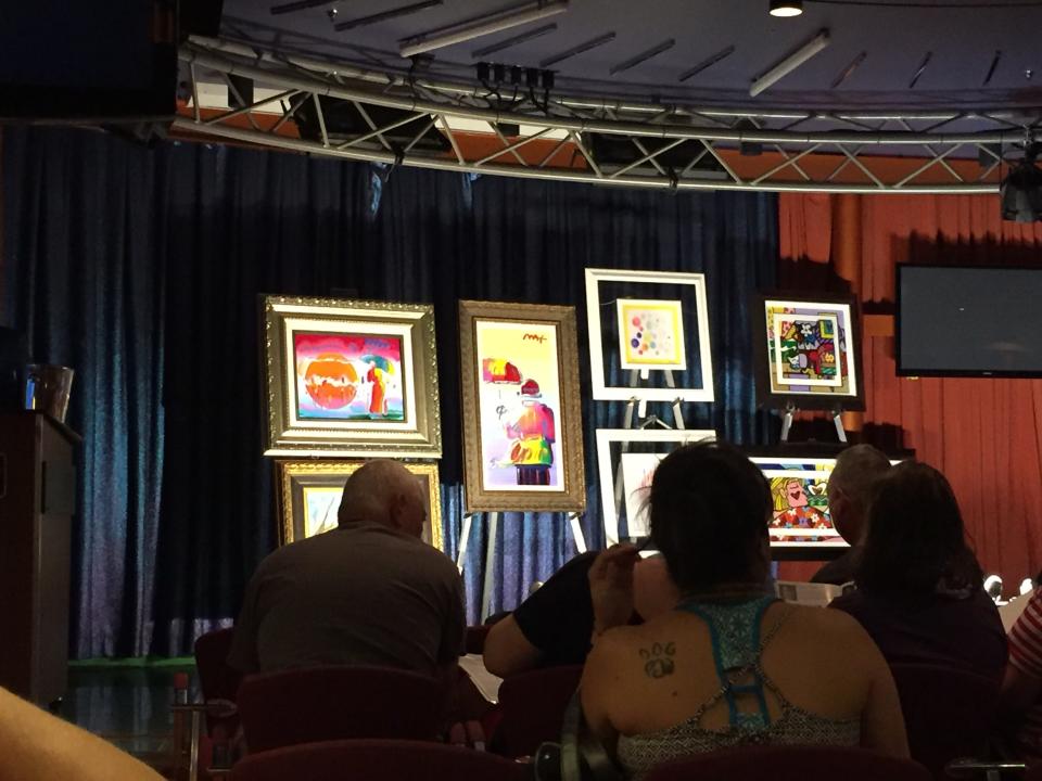 auction room on cruise ship — paintings on display in dark room with people's backs to the camera