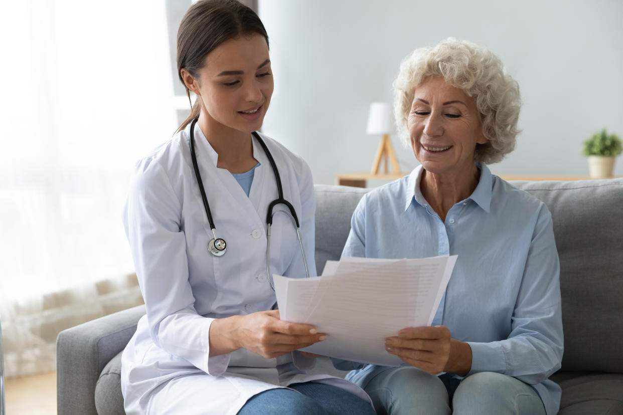 Smiling older woman patient and young nurse doctor holding papers reading health life insurance medical service contract agreement look at test results during homecare visit at home hospital concept