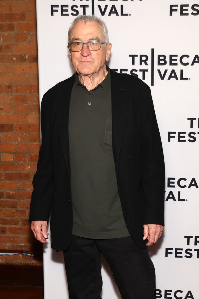 Robert De Niro in a dark blazer and polo shirt at the Tribeca Festival, standing in front of a festival-branded backdrop