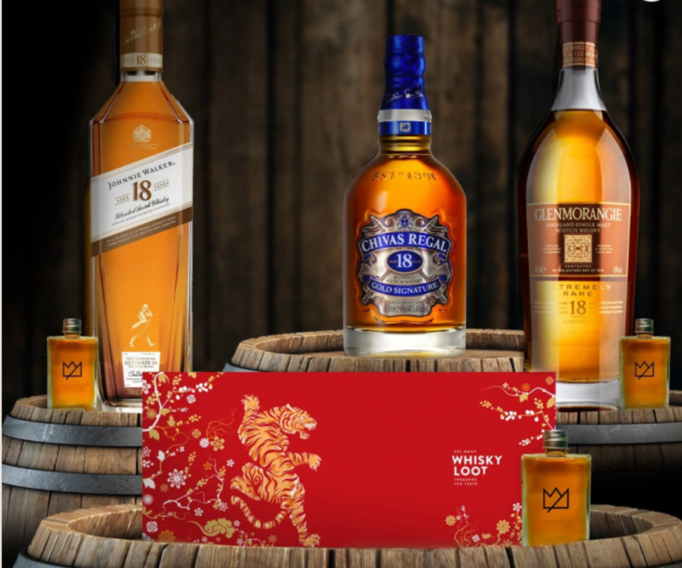 A bottle of Johnnie Walker, Chivas Regal and Glenmorangie whiskeys standing on wooden barrels next to each other against a dark wooden bakground and a red Lunar New Year patterend envelope at the front.