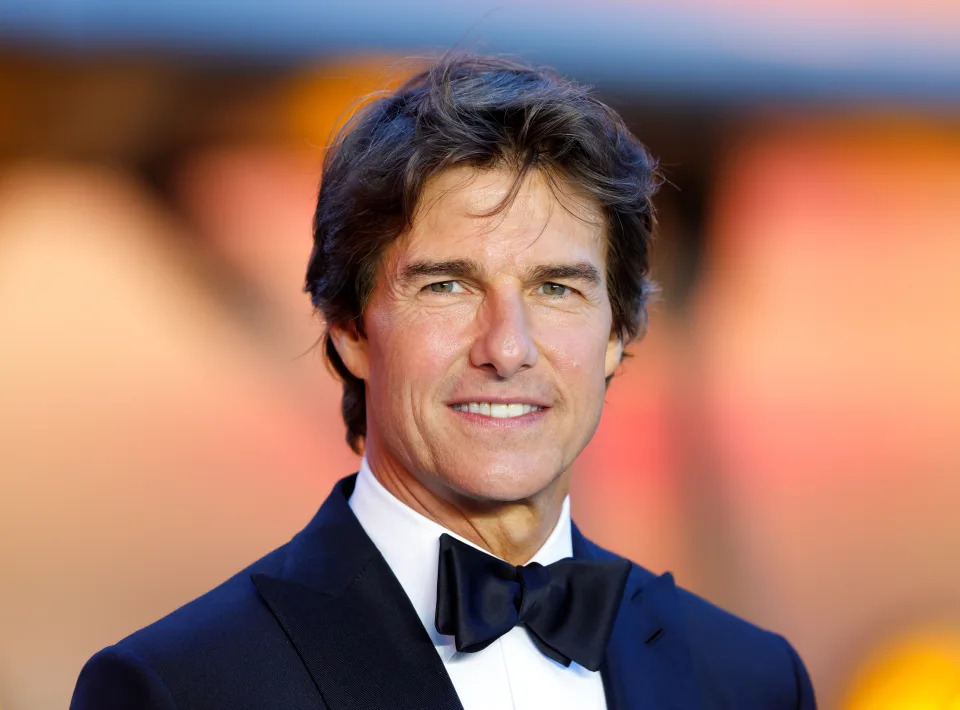 Tom Cruise wearing a classic black tuxedo and bow tie, smiling on the red carpet