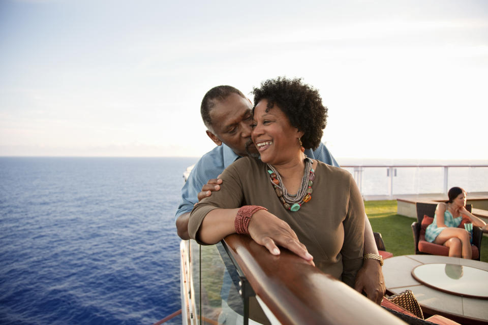 A man lovingly kisses a smiling woman on the cheek on a cruise ship deck, overlooking the ocean. A seated person is in the background