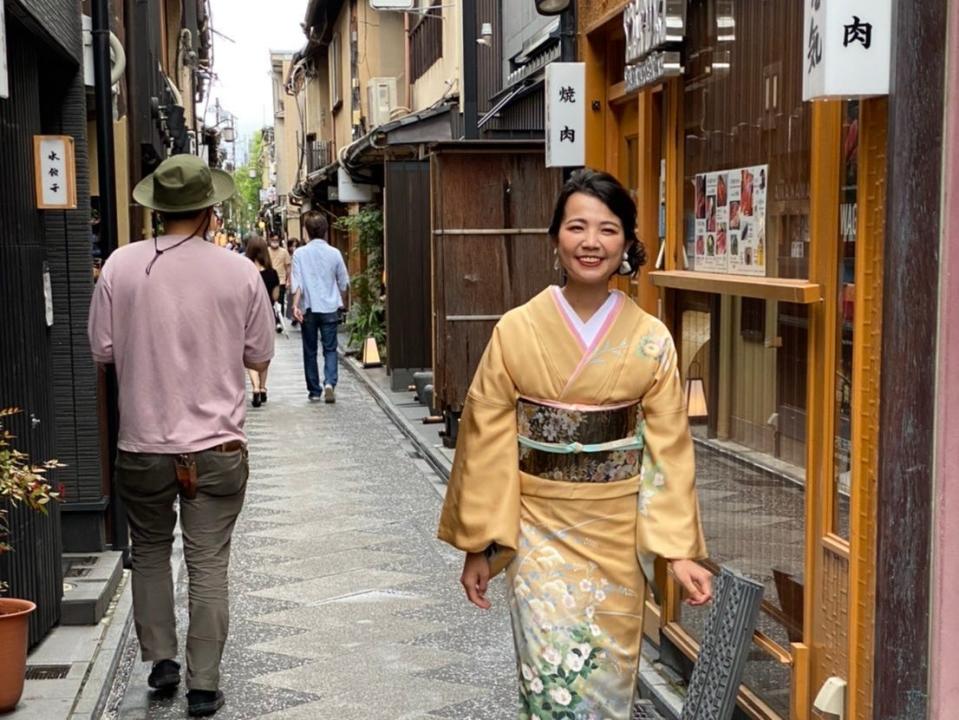 Suganama Natsuki standing in a Japanese alleyway while wearing a traditional-looking yellow dress.