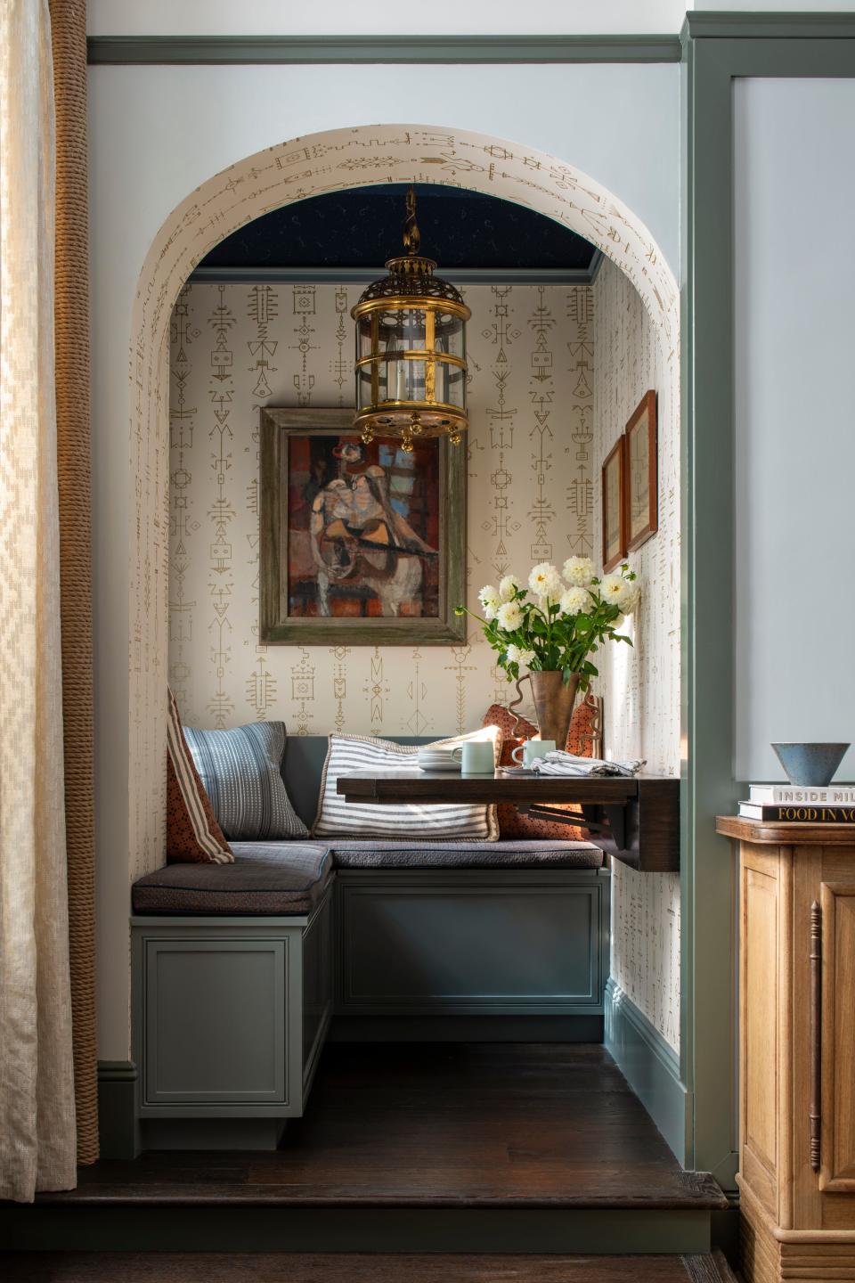 AFTER: The updated dining alcove stars an upholstered banquette, a vintage chandelier, and characterful artworks including a circa 1957 painting by George Ratkai displayed front and center. True to her signature, Louise left no surface bare, not even the arched entrance or the wall behind it, which she outfitted in a Pierre Frey wallpaper.