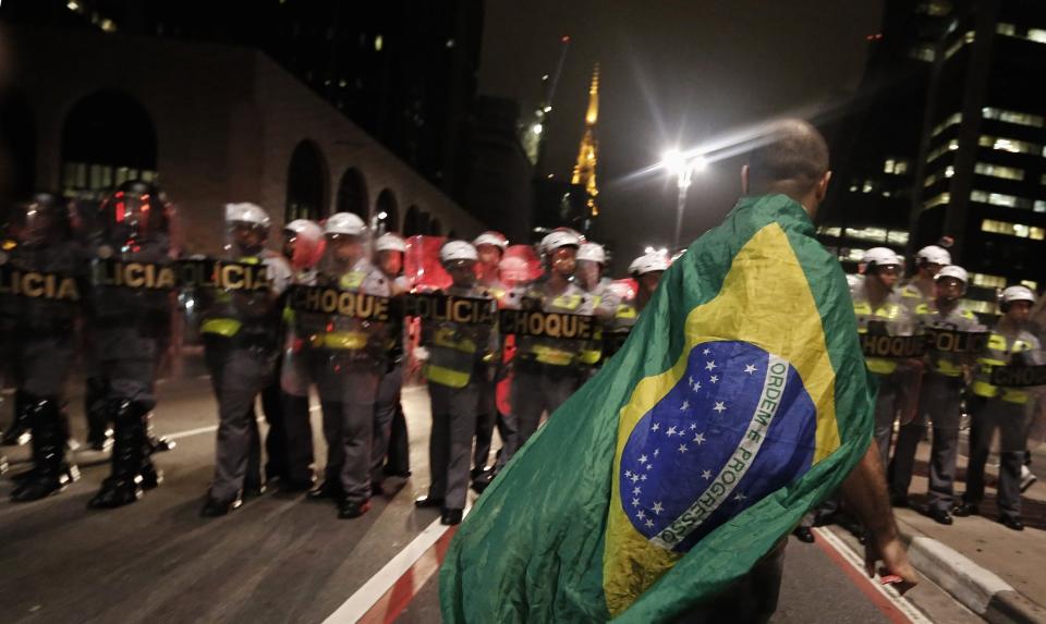 A demonstrator wearing a Brazilian flag walks in front of military policemen during a protest against the 2014 World Cup, in Sao Paulo March 13, 2014. An estimated 1,500 protesters gathered to march the streets of downtown Sao Paulo to show they are against the World Cup and against the costs associated with Brazil's hosting the sporting event. Activists are demanding more money be spent on education, health care, public transportation and to fight crime. REUTERS/Nacho Doce (BRAZIL - Tags: SPORT SOCCER WORLD CUP POLITICS CIVIL UNREST CRIME LAW)