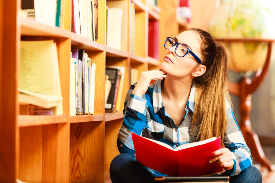 A female college student holding a book while in deep thought in a library.