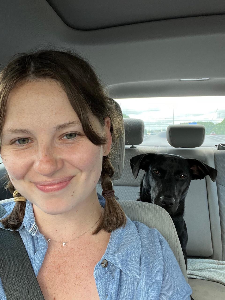 A woman in pigtails smiles in the front seat of a car as a dog looks over her shoulder.