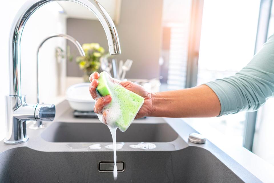 woman's hand squeezing soap out of a green sponge held under running water at kitchen sink