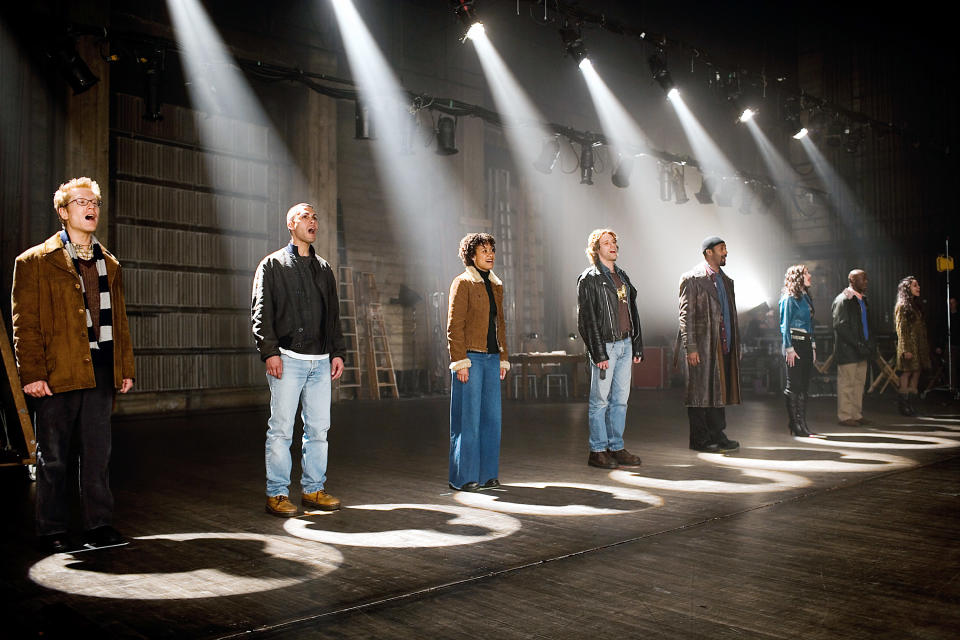 Actors in the 2005 film "Rent" stand in a line on stage, facing forward as a spotlight shines on each of them separately