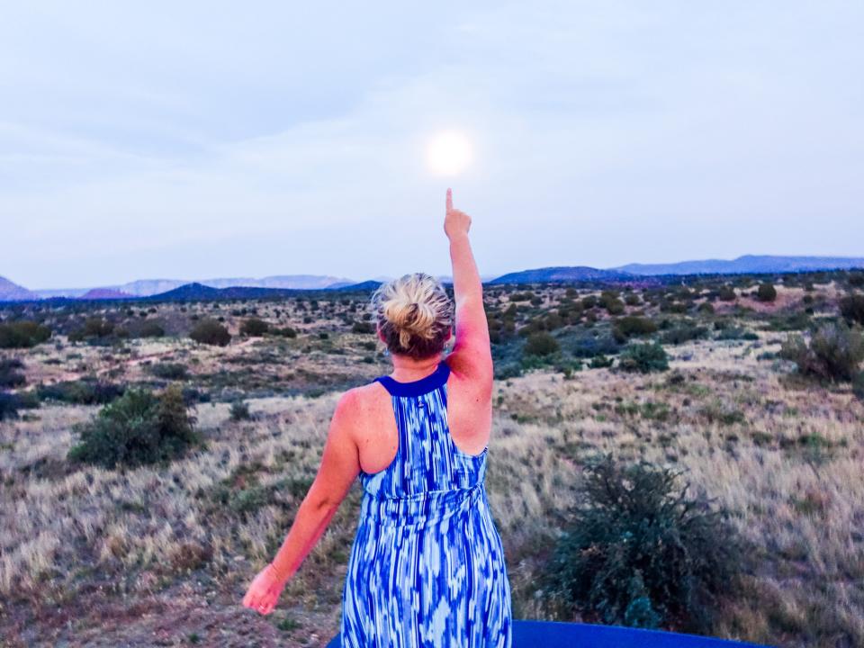 A woman points at the moon over a field in Sedona with cloudy skies.