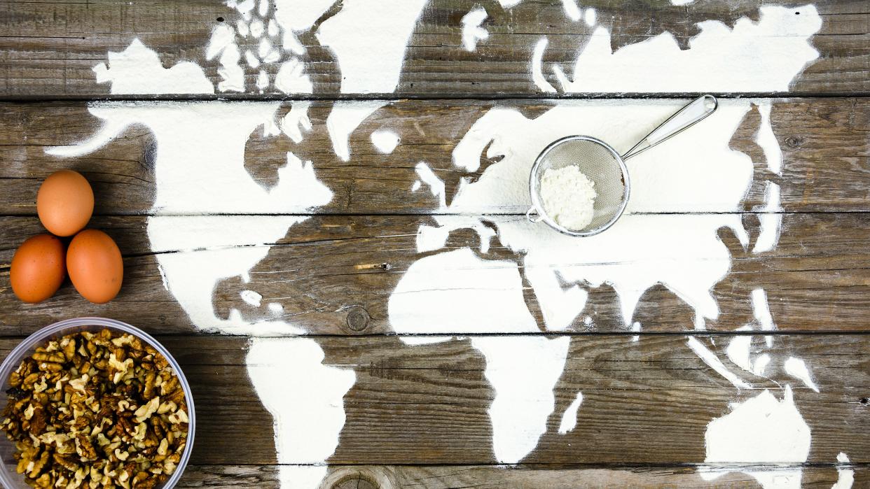  image of world map on a table. three eggs eggs sit to the left of north america. a bowl of grain is left of south america. on top of the border of europe and asia is a sieve with flower 