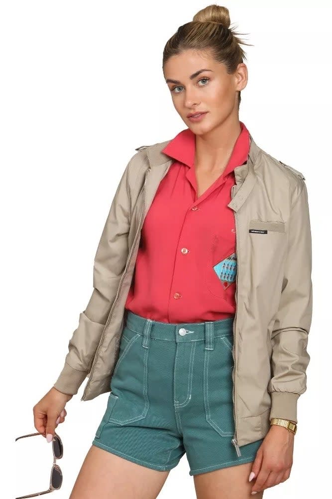 a model in a buttoned-up red shirt, khaki jacket, teal shorts, holding sunglasses
