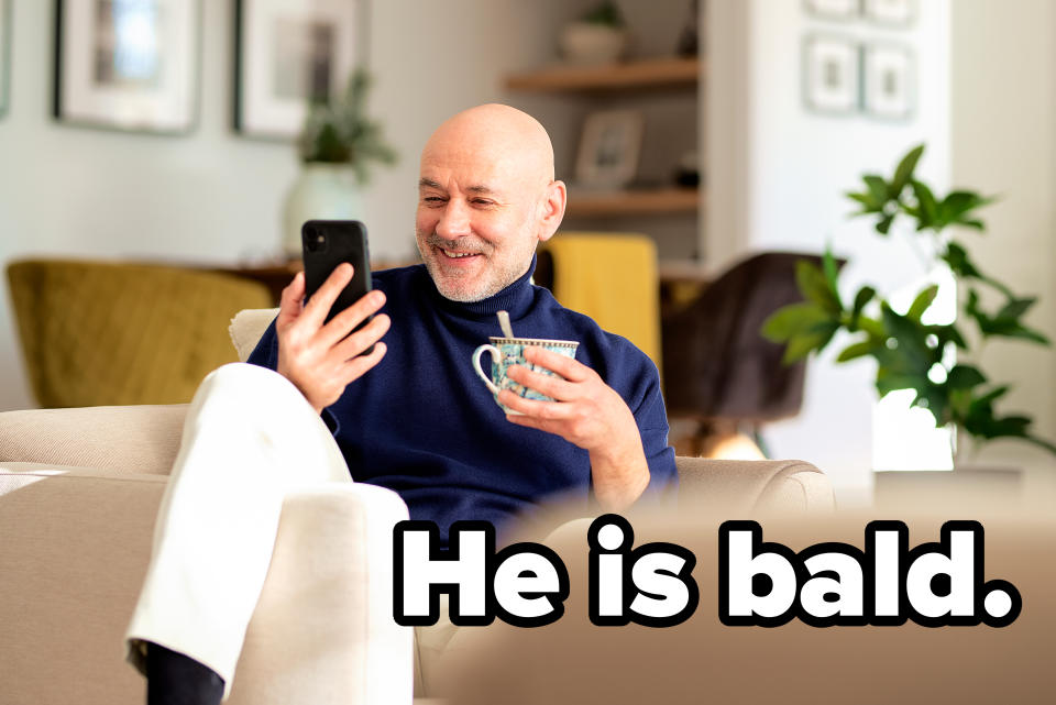 A man sits comfortably on a beige sofa, smiling as he looks at his phone and holds a cup. The background features home decor and greenery