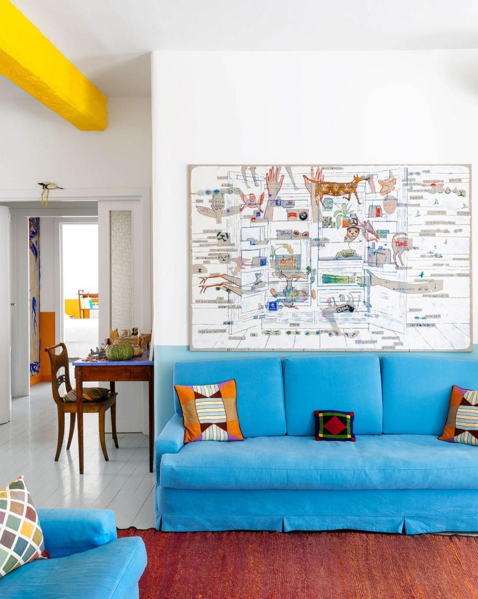 half white and half light blue walls, white wood floor covered with a deep red rug, blue sofa and matching chair both with decorative pillows, small desk and chair, large artwork over sofa, yellow ceiling beam