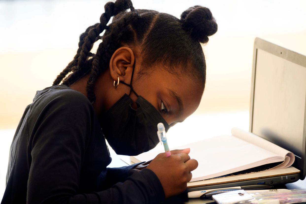 A girl in a face mask painstakingly writes something down at her laptop.