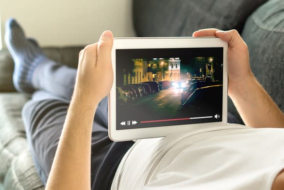 A young man laying on a couch watching a program on a tablet.