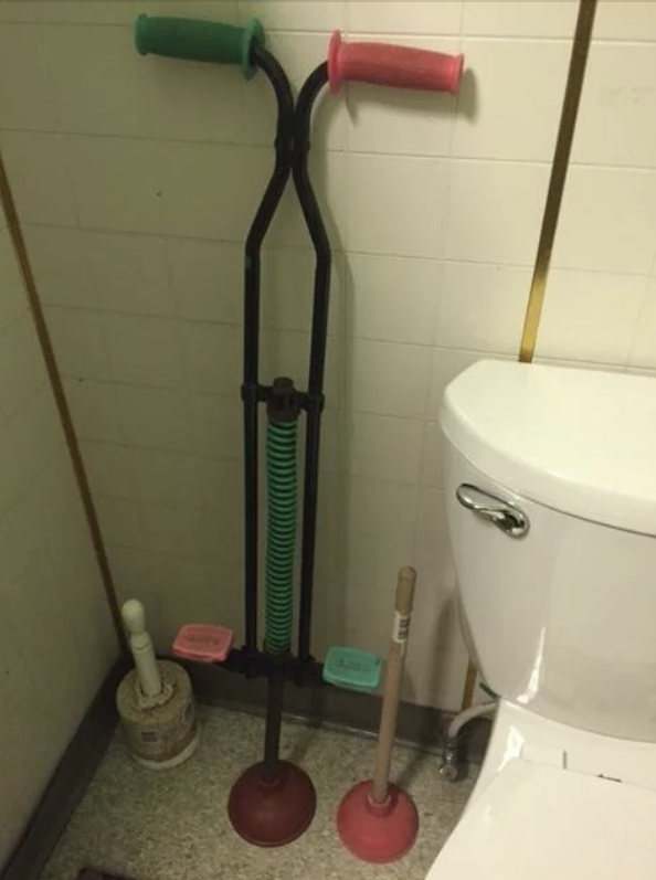 A pogo stick with plunger bases, a regular plunger, and a toilet brush are placed next to a toilet in a bathroom