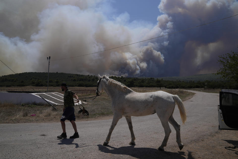 A man evacuates a horse from a stable as smoke plumes from a fire are seen in the background at Pournari village near Athens, Greece, on Tuesday, July 18, 2023. In Greece, where a second heatwave is expected to hit Thursday, three large wildfires burned outside Athens for a second day. Thousands of people evacuated from coastal areas south of the capital returned to their homes Tuesday when a fire finally receded after they spent the night on beaches, hotels and public facilities. (AP Photo/Thanassis Stavrakis)