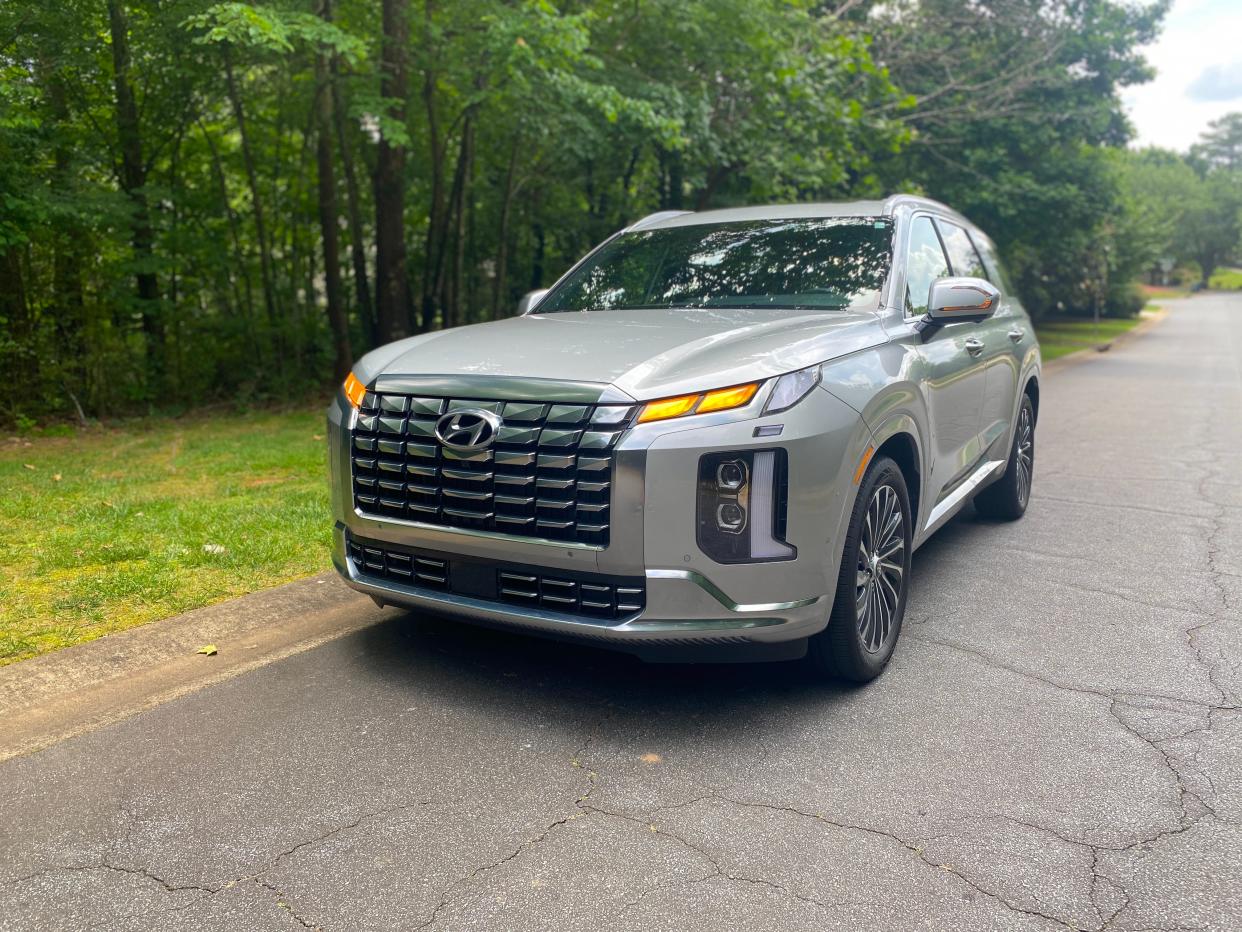 A silver Hyundai Palisade SUV is parked in front of a wooded area with blinkers on.