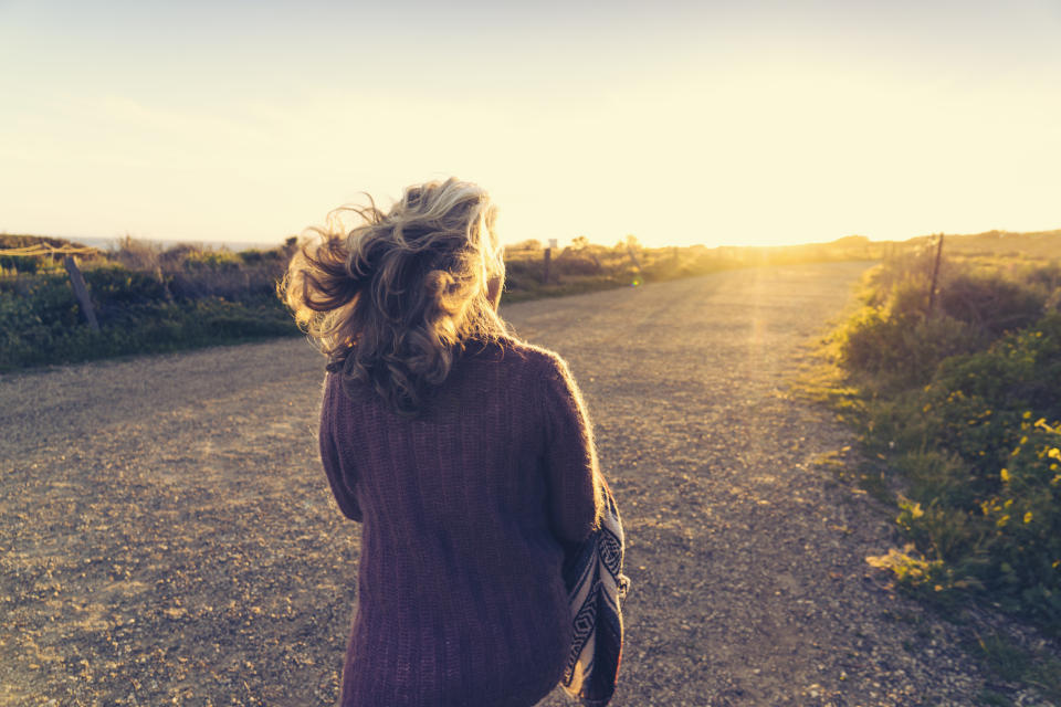 A woman with wavy hair walks along a gravel path during sunset, wrapped in a shawl. The landscape around her is open and serene