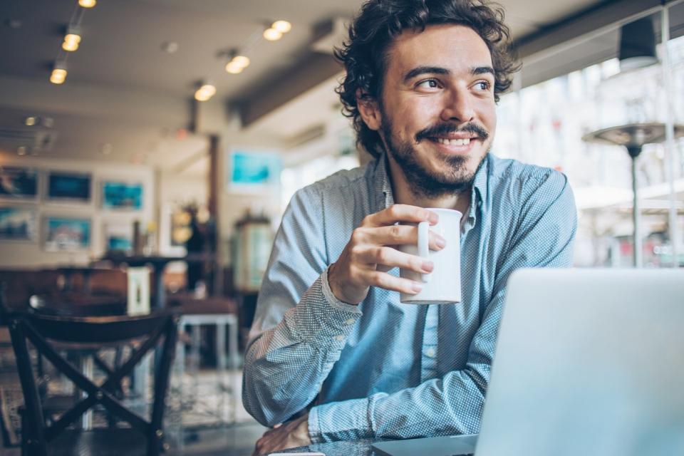 An investor smiles and drinks coffee in front of a laptop in an office.